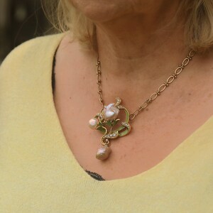 May include: A gold chain necklace with a pendant featuring a pearl flower with green leaves and smaller pearls. The pendant is on a woman's neck.