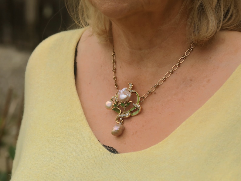 May include: A gold chain necklace with a pendant featuring a pearl flower with green leaves and smaller pearls. The pendant is on a woman's neck.
