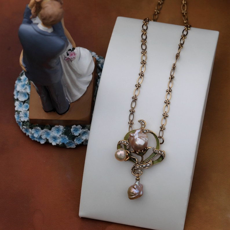 May include: A gold necklace with a delicate chain and a pendant featuring a pink pearl flower with green enamel accents. The pendant is adorned with smaller pearls.