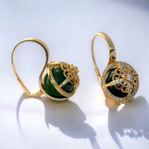 Könnte beinhalten: Ein Paar goldene Ohrringe mit einem grünen Stein, der in einem filigranen Design gefasst ist. Die Ohrringe sind mit kleinen, klaren Steinen verziert.