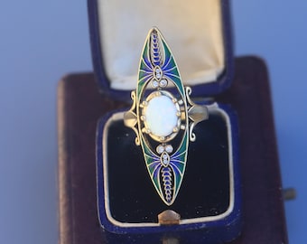 Op de afbeelding: Een Art Deco-stijl ring met een ovale opaal middensteen, omgeven door een gouden filigraan ontwerp. De ring heeft groene en blauwe emaille accenten en kleine diamanten, gepresenteerd in een vintage sieradendoos.
