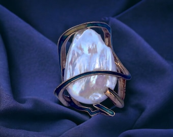 May include: A large, white pearl is set in a gold and blue metal ring. The ring is designed with a series of curved lines that create a cage-like effect around the pearl.