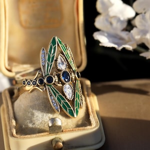 May include: A gold ring featuring a dragonfly design. The dragonfly's wings are green with white and blue accents. The ring is in a small, white box.