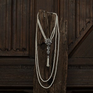 Può includere: Una collana di perle multifilo con un ciondolo in argento con un motivo floreale e un fiocco. La collana è esposta su un pezzo di legno invecchiato.