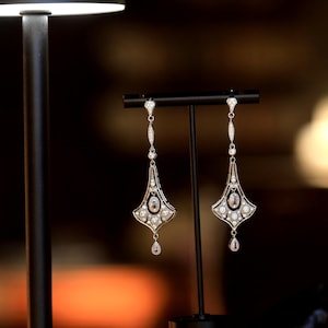 Può includere: Un paio di orecchini pendenti in argento con un diamante a forma di lacrima nella parte inferiore. Gli orecchini sono adornati con piccoli diamanti e hanno un design geometrico.