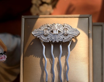 May include: A silver-toned hair comb with an Art Deco design, encrusted with sparkling clear crystals. The comb features a central rectangular crystal and three curved tines. The jewelry is displayed on a neutral-toned surface.