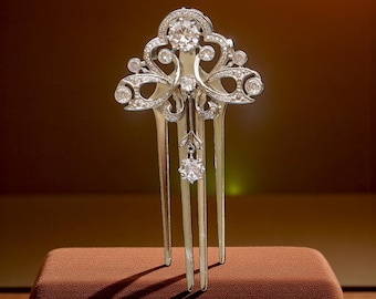 May include: A silver-toned hair comb with an ornate design, featuring multiple clear gemstones. The comb has a central, large round stone and smaller stones throughout the decorative top. Three long prongs extend downward.