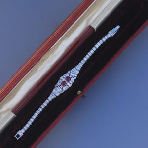 May include: A silver bracelet with a central design featuring two round red gemstones, surrounded by smaller clear stones. The bracelet is in a red velvet lined box.