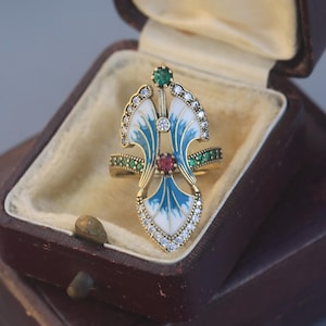 May include: An ornate gold ring with a distinctive design. It features blue and white enamel wings, a central ruby, and emerald and diamond accents. The ring is presented in a vintage brown jewellery box, highlighting its intricate details.
