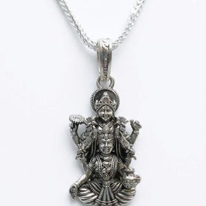May include: A silver pendant featuring a detailed depiction of a deity, suspended from a delicate chain. The pendant showcases intricate carvings of figures, with a detailed design. The chain is a fine, textured link. The background is plain white.