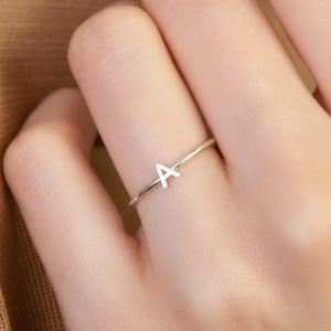 Può includere: Un anello d'argento con la lettera "A" in stile minimalista. L'anello è sottile e delicato, con la lettera centrata sulla fascia. L'anello è indossato su un dito.