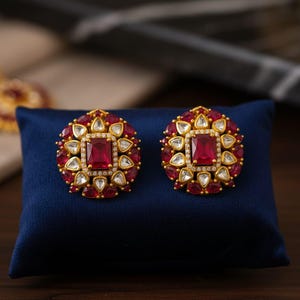 May include: A pair of gold-toned stud earrings with a square, deep red center stone surrounded by smaller red and white gems. The earrings are displayed on a dark blue surface, showcasing their intricate design and vibrant colors.