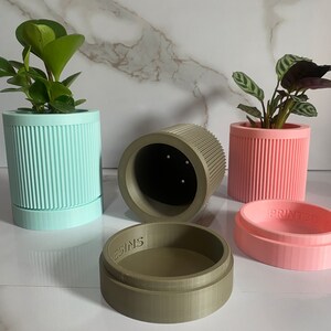 Puede incluir: Cuatro macetas para plantas impresas en 3D en diferentes colores: azul claro, rosa, verde y marr&oacute;n. Las macetas tienen un dise&ntilde;o acanalado y todas son del mismo tama&ntilde;o. Las macetas est&aacute;n sobre una superficie de m&aacute;rmol blanco. El texto "PRINTED" es visible en la maceta rosa y el texto "DESIGNS" es visible en la maceta verde.