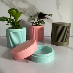 Puede incluir: Tres macetas cil&iacute;ndricas con texturas acanaladas en verde, rosa y marr&oacute;n. Las macetas est&aacute;n hechas de resina y tienen un dise&ntilde;o minimalista. Una maceta tiene una tapa rosa.