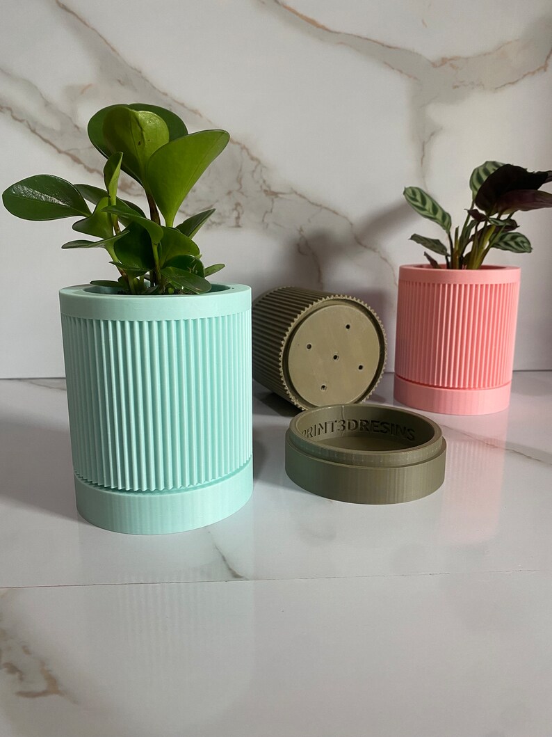 Puede incluir: Tres macetas para plantas impresas en 3D, una verde claro, una rosa y una verde oscuro. La maceta verde claro tiene una planta dentro. La maceta verde oscuro tiene el texto "PRINT3DRESINS" en el lateral.