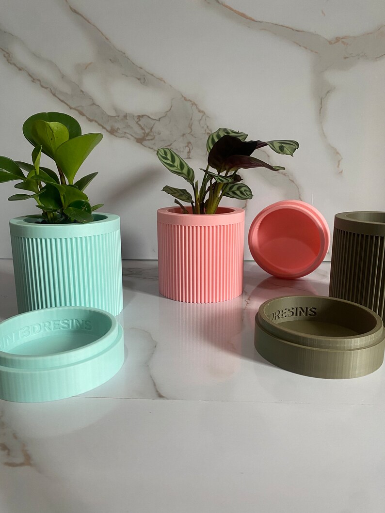 Puede incluir: Tres macetas cil&iacute;ndricas con tapas, una en azul claro, una en rosa y otra en verde oliva. Cada maceta tiene un dise&ntilde;o acanalado y la palabra "3DResins" impresa en la parte inferior.