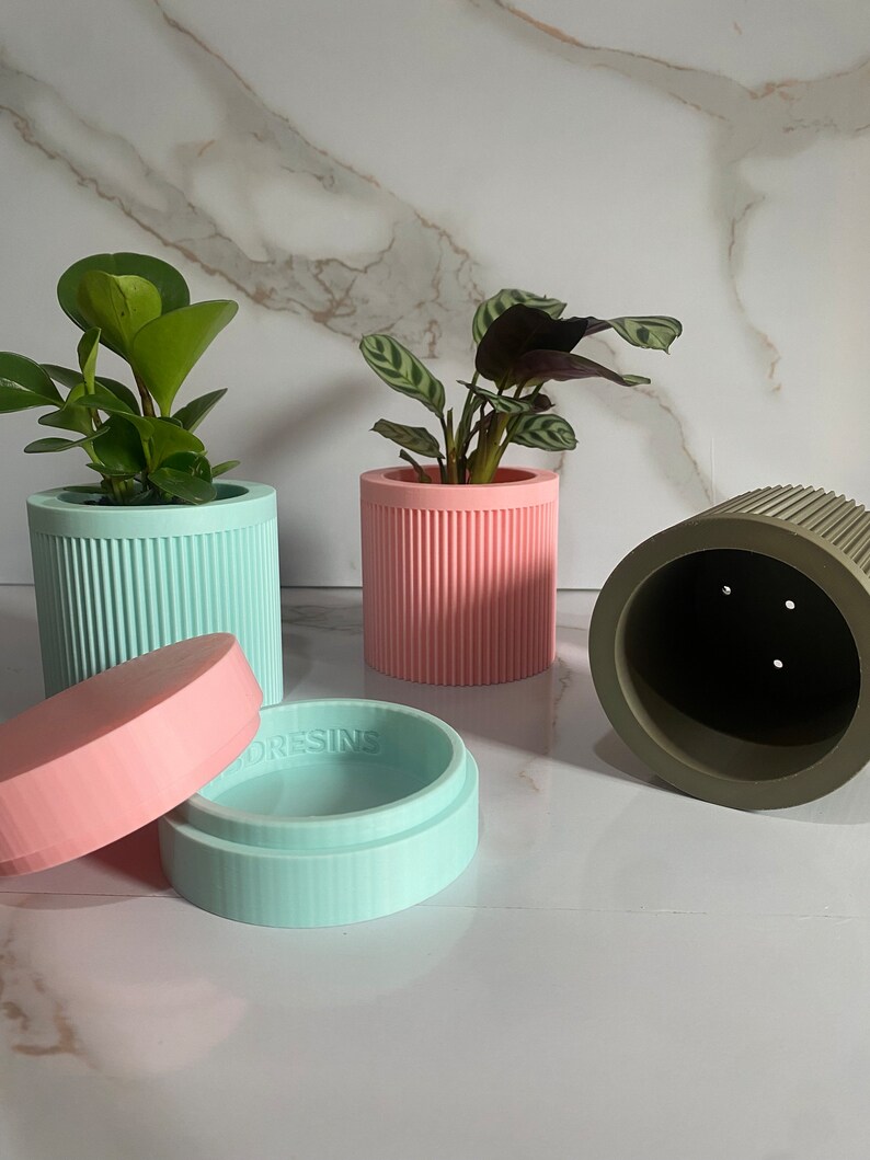 Puede incluir: Tres macetas para plantas, una rosa, una azul claro y una verde. Las macetas rosa y azul claro tienen una textura acanalada. La maceta verde tiene agujeros de drenaje. La maceta azul claro tiene una tapa con la palabra "ORESINS" impresa en ella. Las macetas est&aacute;n sobre una superficie de m&aacute;rmol blanco.