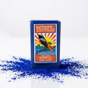 Può includere: Un contenitore rettangolare blu di pigmento Blu Oltremare Sunshine. L'etichetta presenta un'illustrazione di uccello con un disegno di sole nascente. Il pigmento è sparso intorno al contenitore.