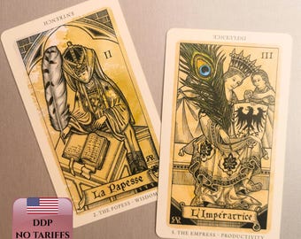 Op de afbeelding: Twee tarotkaarten met gedetailleerde illustraties. Eén kaart toont een figuur die met een veer schrijft, getiteld "La Papesse" en "The Popess - Wisdom". De andere kaart toont een figuur met een pauwenveer, getiteld "L'Impératrice" en "The Empress - Productivity."