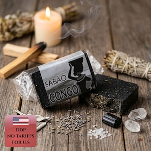 Peut inclure: Deux pains de savon noir rectangulaires, dont l'un est enveloppé d'une étiquette en papier portant le texte SABÃO CONGO et la silhouette d'une personne portant un panier sur la tête. Le savon est texturé avec des herbes et des particules sombres.