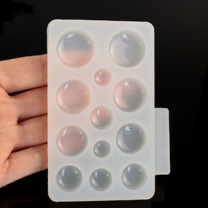 Op de afbeelding: Een doorzichtige siliconen mal met meerdere cirkelvormige holtes van verschillende groottes. De holtes zijn gerangschikt in een rechthoekig patroon. Sommige bollen hebben een roze tint, andere zijn grijs. De mal is gelabeld "HobbyMagi".