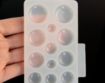 May include: A clear silicone mold with multiple circular cavities of varying sizes. The cavities are arranged in a rectangular pattern. Some spheres have a pinkish hue, while others are gray. The mold is labeled "HobbyMagi"