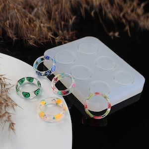 May include: Six clear silicone moulds for making resin rings. Four rings are shown, each with a different colourful design.