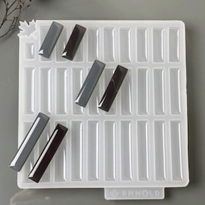 Könnte beinhalten: Eine weiße Silikonform mit rechteckigen Hohlräumen, die mehrere dunkelgraue und schwarze Harzstücke enthält. Der Text "SILICONE MOLD" steht oben. Zusätzlicher Text lautet "- 30 in 1 - Für Harz - Schmuckherstellung."