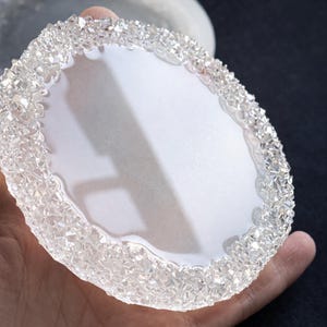 Può includere: Un vassoio decorativo ovale trasparente con un bordo strutturato simile a cristalli. Il vassoio ha un interno bianco ed è tenuto in una mano. Lo sfondo è scuro.