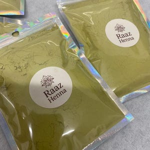 May include: Close-up of several sealed pouches containing green powder, possibly henna. Each pouch has a silver, iridescent top and a white circular label with the text "Raaz Henna". The pouches are arranged closely together, suggesting a product display.