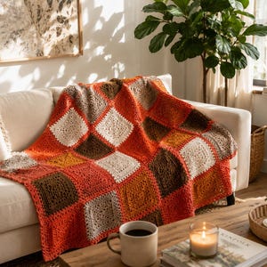 Può includere: Una coperta all'uncinetto fatta a mano con un motivo ripetitivo di quadrati nei toni dell'arancione bruciato, crema, marrone scuro e beige. Il tessuto è composto da quadrati di filato uniti insieme per formare un grande plaid rettangolare.
