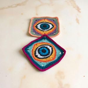 May include: Two handmade crochet squares with an eye design. The squares feature a central blue eye surrounded by concentric circles of orange, yellow, and white. The edges are in shades of pink, teal, and purple. The squares are approximately 10 cm by 10 cm.