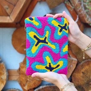 Könnte beinhalten: Eine rechteckige, leuchtend rosa Clutch mit einem farbenfrohen, abstrakten Design. Das Muster zeigt blaue, gelbe und dunkelblaue Formen. Die Clutch hat einen Reißverschluss und wird von einer Person gehalten.