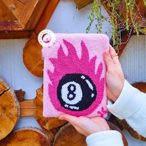 Könnte beinhalten: Rosa Filztasche mit einem schwarzen und weißen 8-Ball-Design. Der 8-Ball ist von rosa Flammen umgeben. Die Tasche wird von einer Hand gehalten.