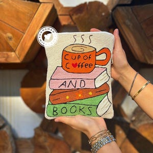 May include: Cream-coloured beaded clutch bag with a design of a coffee cup and a stack of books. The orange cup says "Cup of Coffee" and the books say "And Books". The bag is held by a person.
