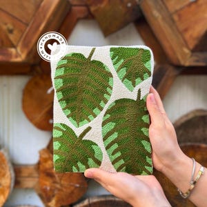 Könnte beinhalten: Eine weiße Clutch-Tasche mit grünem Blatt-Muster. Die Tasche ist aus Perlen gefertigt und trägt den Schriftzug "Handmade with Love" auf einem Etikett.