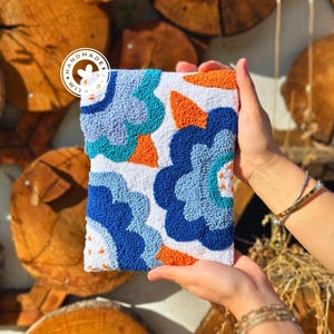 Puede incluir: Una bolsa blanca con un diseño floral colorido. La bolsa está hecha con una técnica de aguja de punzón y presenta flores azules, naranjas y turquesas. La bolsa tiene una etiqueta hecha a mano que dice "LIM Handmade".