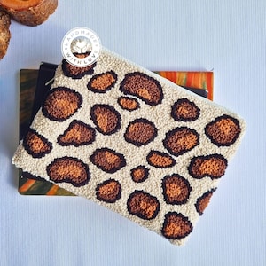 Puede incluir: Un bolso de mano blanco con un diseño de estampado de leopardo marrón y naranja. El bolso está hecho con un tejido esponjoso y texturizado y tiene una pequeña etiqueta redonda que dice "Handmade with Love".