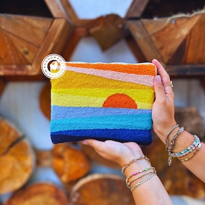 May include: A handmade rectangular clutch bag with a sunset design. The artwork shows a vibrant sunset over the ocean, with layers of orange, yellow, blue, and navy. The clutch is held in front of a wooden background. The text "HANDMADE WITH LOVE" is on the clutch.