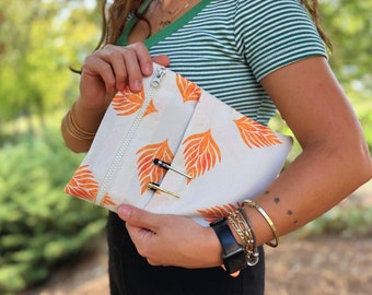 Könnte beinhalten: Eine weiße Canvas-Clutch mit Reißverschluss und einem orangefarbenen Blattmotiv. Die Clutch wird von einer Person gehalten und enthält zwei silberne Stifte.