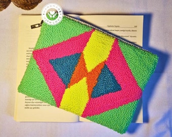 May include: A colorful geometric patterned pouch with a zipper closure. The pouch is made of a textured fabric with a green, yellow, pink, blue, and orange color scheme. The pouch is sitting on top of a book.