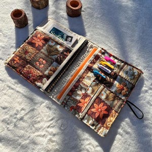 Pode incluir: Uma capa de livro de tecido acolchoado com um design de patchwork floral em tons de castanho, laranja e creme. A capa aberta revela um caderno, canetas e um bolso com um cartão. A capa tem uma alça preta.