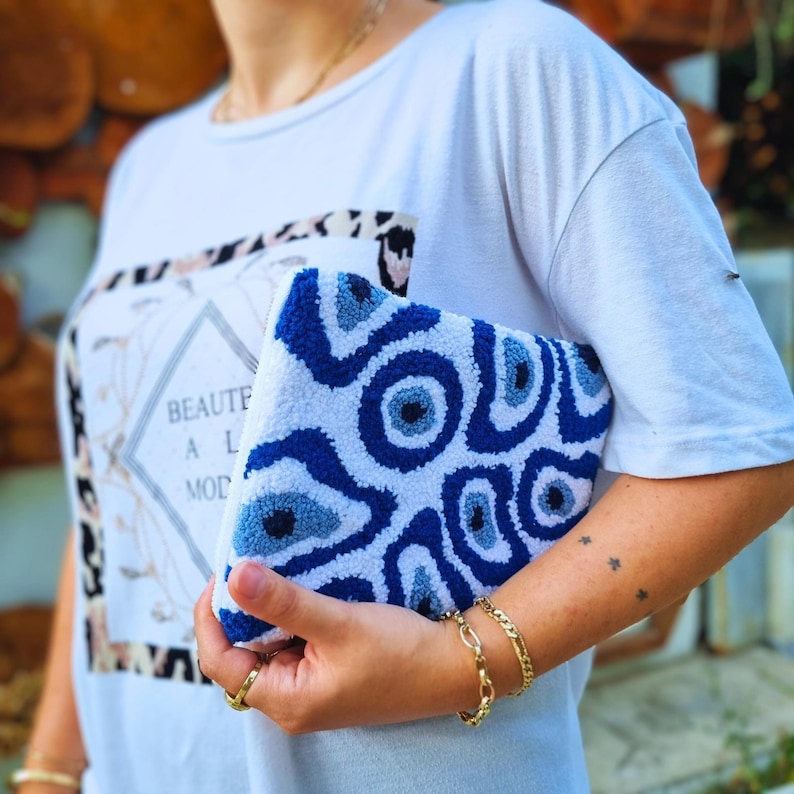 May include: A white clutch bag with a blue and white evil eye pattern. The bag is made of a soft, plush material and has a zipper closure.