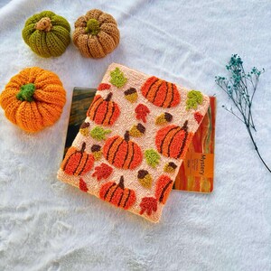 May include: A close-up of a handmade needle felted pumpkin pouch with a light pink background and a pattern of orange pumpkins, brown acorns, and green leaves. The pouch is sitting on top of a brown and orange notebook with the word "Mystery" printed on the cover.