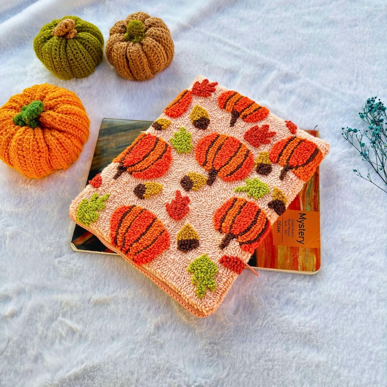 May include: A pink fabric pouch with a zipper closure features a repeating pattern of orange pumpkins, green leaves, and brown acorns. The pouch is lying on top of a notebook with the word "Mystery" printed on the cover.