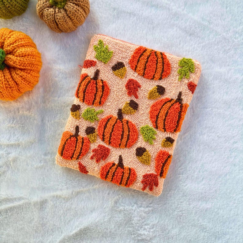 May include: A peach-colored pouch with a repeating pattern of orange pumpkins, brown acorns, and green leaves. The pouch is made of a soft, textured fabric.