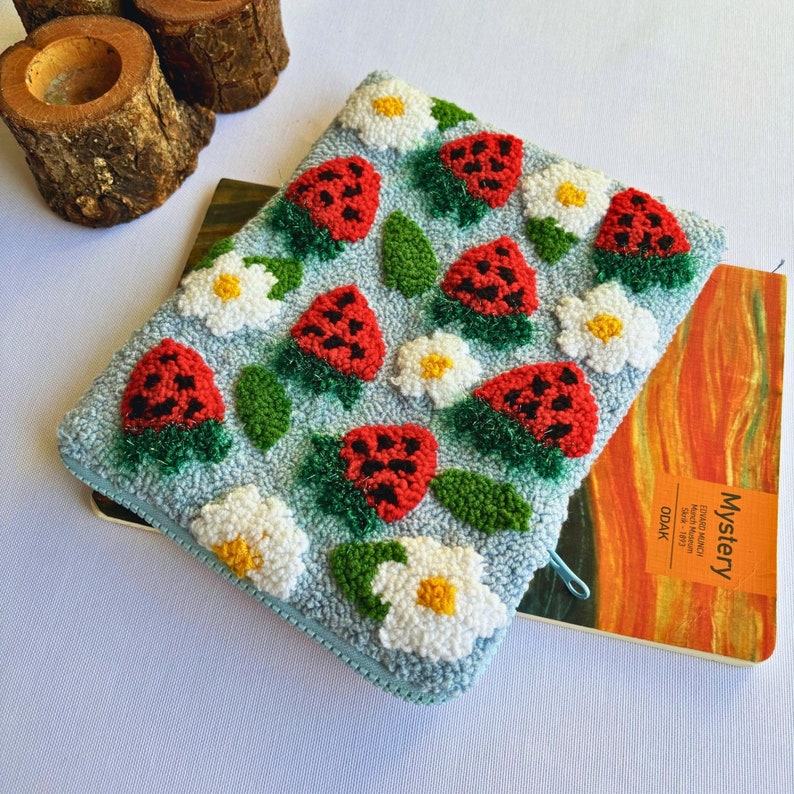 Peut inclure: Une pochette bleu clair avec une fermeture &eacute;clair pr&eacute;sente un motif r&eacute;p&eacute;t&eacute; de fraises rouges et de marguerites blanches. La pochette est faite d'un tissu textur&eacute; et duveteux.