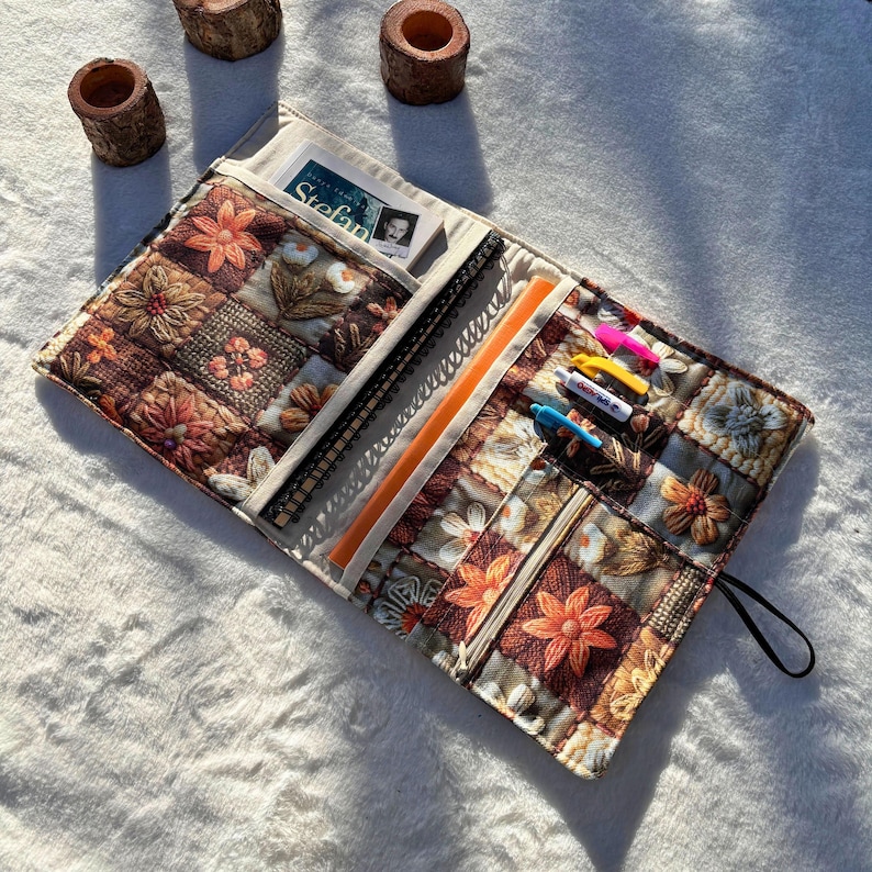 May include: A quilted fabric book cover with a floral patchwork design in shades of brown, orange, and cream. The cover is open, revealing a notebook, pens, and a pocket with a card. The cover has a black strap.