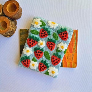 Peut inclure: Une pochette en tissu bleu avec une fermeture &eacute;clair blanche. La pochette est d&eacute;cor&eacute;e de fraises brod&eacute;es et de fleurs blanches. La pochette est pos&eacute;e sur un carnet jaune et orange avec le mot "Mystery" imprim&eacute; sur la couverture.