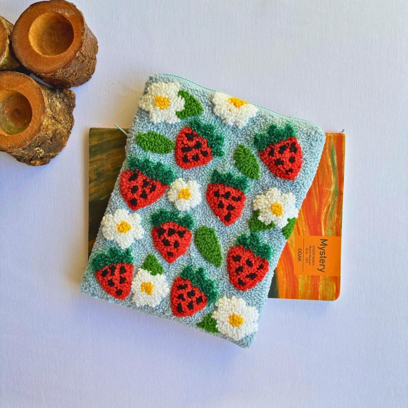 Peut inclure: Une pochette en tissu bleu avec une fermeture &eacute;clair blanche. La pochette est d&eacute;cor&eacute;e de fraises brod&eacute;es et de fleurs blanches. La pochette est pos&eacute;e sur un carnet jaune et orange avec le mot "Mystery" imprim&eacute; sur la couverture.